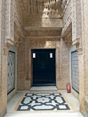 An intricately carved stone doorway with detailed geometric patterns surrounds an entrance to a dimly lit interior. The ornate architecture features elaborate arabesques and calligraphy, with a small fire extinguisher placed discretely to the side on the marble floor which has a black and white geometric design.