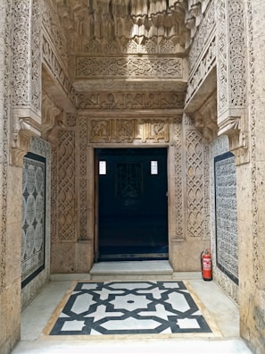 An intricately carved stone doorway with detailed geometric patterns surrounds an entrance to a dimly lit interior. The ornate architecture features elaborate arabesques and calligraphy, with a small fire extinguisher placed discretely to the side on the marble floor which has a black and white geometric design.