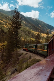 A scenic mountain landscape with a vintage green train traveling along a railway track surrounded by dense forests. The mountains are lush with trees, some exhibiting autumn colors of yellow and orange, under a partly cloudy sky.