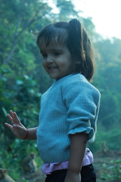 A young child is smiling in a natural setting, wearing a light blue sweater with a pink shirt underneath. The background features a lush green forest with some monkeys visible.