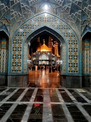A beautifully ornate mosque entrance with intricate tile patterns showcasing geometric designs in blue, yellow, and white colors. The interior displays an illuminated golden dome and minarets. A group of people is gathered under the archway, some dressed in traditional attire.