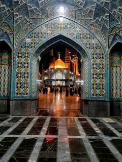 A beautifully ornate mosque entrance with intricate tile patterns showcasing geometric designs in blue, yellow, and white colors. The interior displays an illuminated golden dome and minarets. A group of people is gathered under the archway, some dressed in traditional attire.