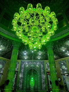 An ornate interior with a large, intricate chandelier glowing in green, surrounded by decorative columns and arches. The walls are adorned with elaborate Islamic patterns and calligraphy, creating a rich and detailed visual atmosphere.