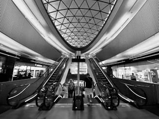 A modern architectural interior with two escalators leading upwards, framed by a geometric patterned ceiling. The space is lined with metallic panels and illuminated by ambient lighting. Several people are seen standing at the base of the escalators, likely in a shopping or transit center environment.