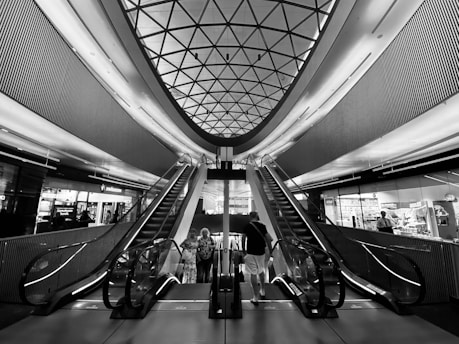 A modern architectural interior with two escalators leading upwards, framed by a geometric patterned ceiling. The space is lined with metallic panels and illuminated by ambient lighting. Several people are seen standing at the base of the escalators, likely in a shopping or transit center environment.