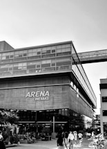 A modern building with glass windows houses the Arena Cinemas. People are walking around and sitting in an outdoor area, with a few trees and plants enhancing the urban setting.