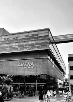 A modern building with glass windows houses the Arena Cinemas. People are walking around and sitting in an outdoor area, with a few trees and plants enhancing the urban setting.