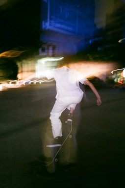 An abstract, motion-blurred scene of a person skateboarding at night. The skater is wearing white clothing, and their movement creates a dynamic, ghost-like effect. Bright light trails and dark surroundings suggest nighttime city photography.