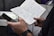 A close-up of a person holding an open notebook filled with handwritten notes and a pen in one hand, and a smartphone in the other. The setting appears to be an indoor environment with some cables and a vent visible in the background.