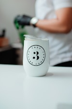 A white tumbler with the logo of 'TRES ANA COFFEE COMPANY' is placed on a smooth, white surface. In the background, a person wearing a white shirt and a watch on their wrist is visible, with a gloved hand holding a green object.