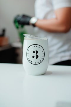 A white tumbler with the logo of 'TRES ANA COFFEE COMPANY' is placed on a smooth, white surface. In the background, a person wearing a white shirt and a watch on their wrist is visible, with a gloved hand holding a green object.