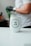 A white tumbler with the logo of 'TRES ANA COFFEE COMPANY' is placed on a smooth, white surface. In the background, a person wearing a white shirt and a watch on their wrist is visible, with a gloved hand holding a green object.