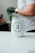 A white tumbler with the logo of 'TRES ANA COFFEE COMPANY' is placed on a smooth, white surface. In the background, a person wearing a white shirt and a watch on their wrist is visible, with a gloved hand holding a green object.