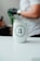 A white tumbler with the logo of 'TRES ANA COFFEE COMPANY' is placed on a smooth, white surface. In the background, a person wearing a white shirt and a watch on their wrist is visible, with a gloved hand holding a green object.