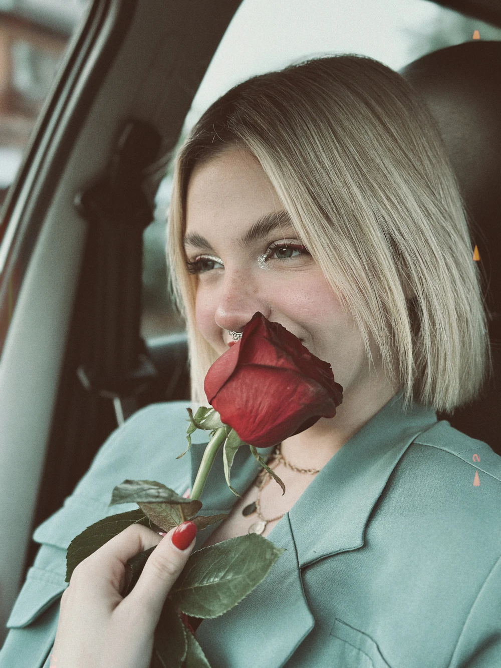 Margaux in the passenger seat of a car holding a red rose to her lips.