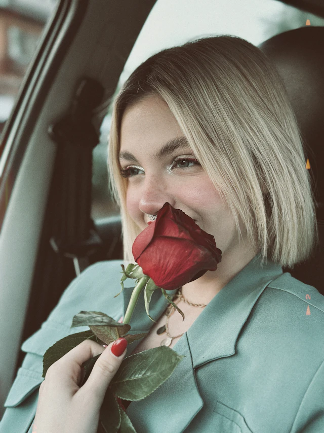 Margaux in the passenger seat of a car holding a red rose to her lips.