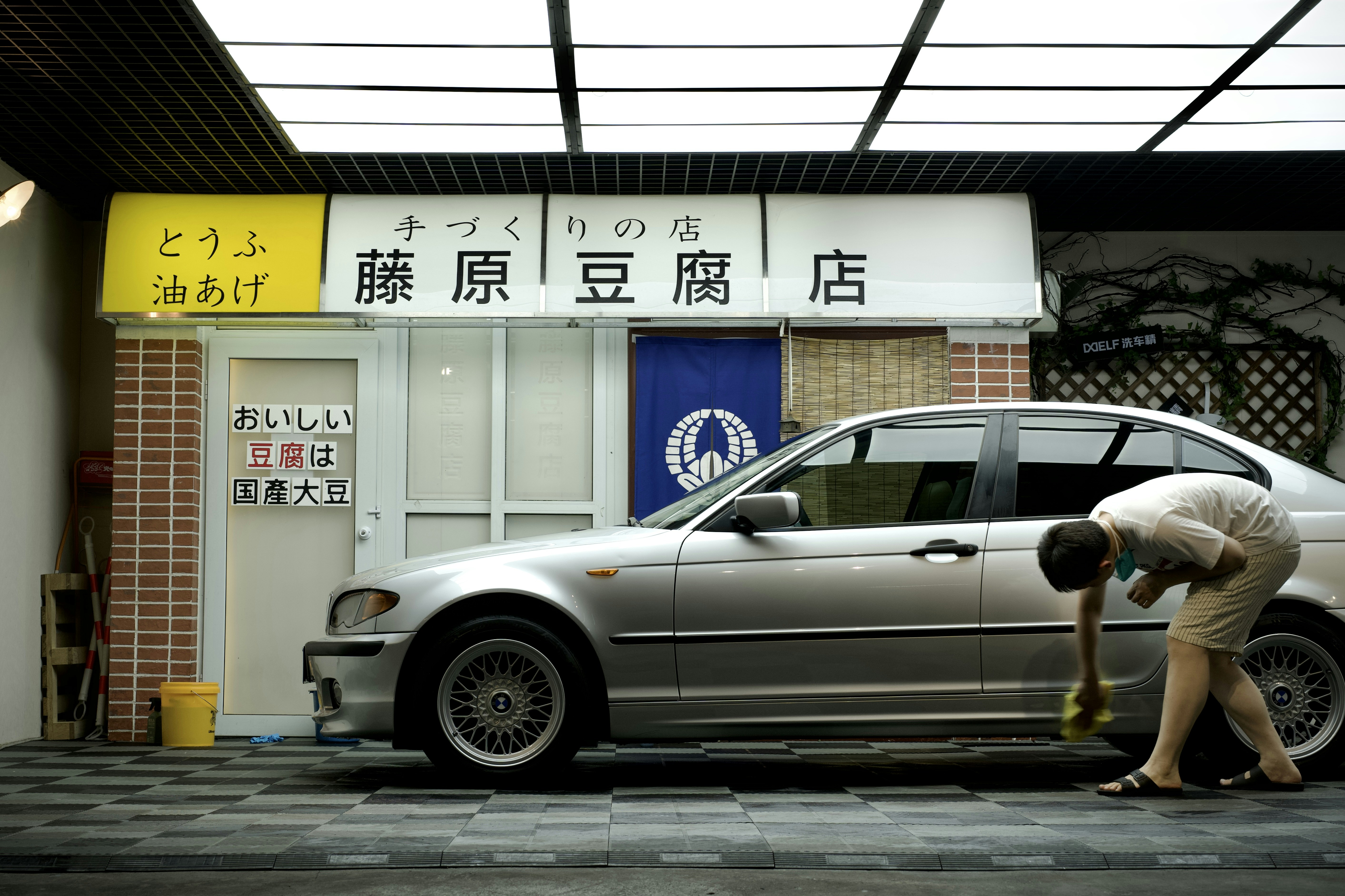 A person is cleaning a silver car parked in front of a tofu shop. The shop has a yellow and white sign with Japanese writing and a blue curtain with a white emblem. The exterior includes a brick pillar and a tiled surface.