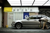 A person is cleaning a silver car parked in front of a tofu shop. The shop has a yellow and white sign with Japanese writing and a blue curtain with a white emblem. The exterior includes a brick pillar and a tiled surface.