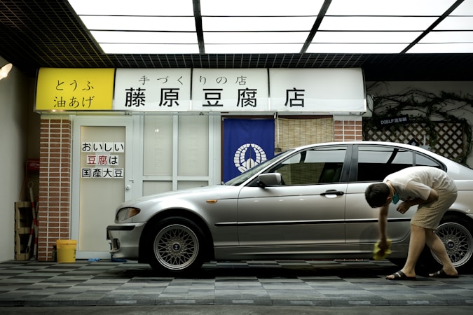 A person is cleaning a silver car parked in front of a tofu shop. The shop has a yellow and white sign with Japanese writing and a blue curtain with a white emblem. The exterior includes a brick pillar and a tiled surface.