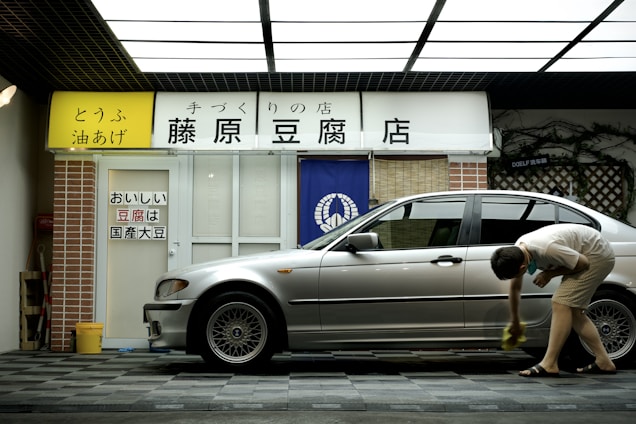 A person is cleaning a silver car parked in front of a tofu shop. The shop has a yellow and white sign with Japanese writing and a blue curtain with a white emblem. The exterior includes a brick pillar and a tiled surface.