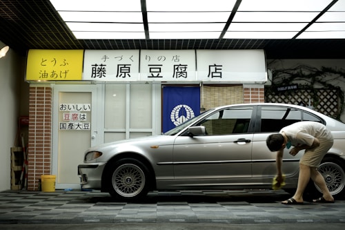 A person is cleaning a silver car parked in front of a tofu shop. The shop has a yellow and white sign with Japanese writing and a blue curtain with a white emblem. The exterior includes a brick pillar and a tiled surface.