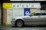 A silver car is parked in front of a tofu shop, which has a white and blue entrance with Japanese signage. There is a brick wall on the left side and a yellow bucket on the ground nearby.