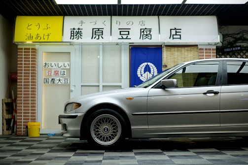 A silver car is parked in front of a tofu shop, which has a white and blue entrance with Japanese signage. There is a brick wall on the left side and a yellow bucket on the ground nearby.
