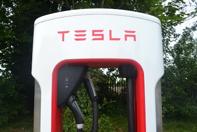 Close-up view of a Tesla electric vehicle charging station placed amidst a backdrop of green foliage. The station has a prominent red and white design, with the Tesla logo displayed at the top.
