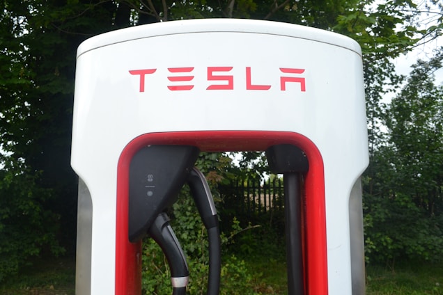 Close-up view of a Tesla electric vehicle charging station placed amidst a backdrop of green foliage. The station has a prominent red and white design, with the Tesla logo displayed at the top.