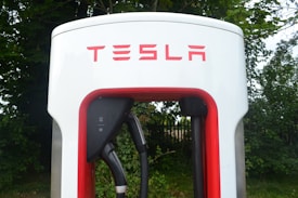 Close-up view of a Tesla electric vehicle charging station placed amidst a backdrop of green foliage. The station has a prominent red and white design, with the Tesla logo displayed at the top.