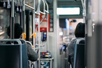 The inside of a bus is captured, showing two passengers seated. The interior features metal handrails, fabric-covered seats, and overhead handles for standing passengers. The focus is on the back of the bus seats and parts of the people seated, with signage and a payment terminal visible near the entrance.