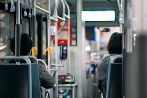 The inside of a bus is captured, showing two passengers seated. The interior features metal handrails, fabric-covered seats, and overhead handles for standing passengers. The focus is on the back of the bus seats and parts of the people seated, with signage and a payment terminal visible near the entrance.