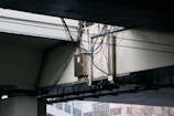 A bundle of cables is attached to a metal conduit running under a large concrete overpass. The overpass structure is weathered, and some urban buildings are visible in the background.