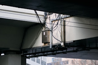 A bundle of cables is attached to a metal conduit running under a large concrete overpass. The overpass structure is weathered, and some urban buildings are visible in the background.
