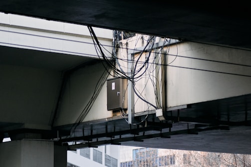 A bundle of cables is attached to a metal conduit running under a large concrete overpass. The overpass structure is weathered, and some urban buildings are visible in the background.