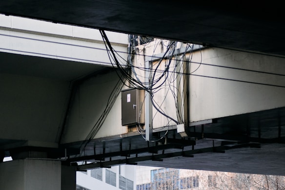 A bundle of cables is attached to a metal conduit running under a large concrete overpass. The overpass structure is weathered, and some urban buildings are visible in the background.