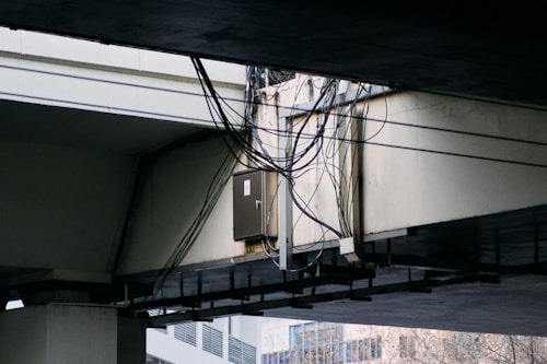 A bundle of cables is attached to a metal conduit running under a large concrete overpass. The overpass structure is weathered, and some urban buildings are visible in the background.