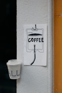 A drawing is pinned to a wall depicting an IV bag labeled with '300ml' and 'COFFEE' inside it. Below the drawing is an upside-down disposable coffee cup attached to the wall. The scene is set against a white textured surface and a partial view of an orange frame on the right.