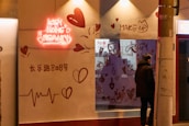 A storefront with a neon sign reading 'ICEY HOME BEAUTY' is illuminated. The wall features heart designs and various beauty-related doodles such as makeup items. A person stands outside the shop, leaning against a streetlight pole. The scene appears to be in an urban setting during the evening.