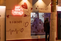 A storefront with a neon sign reading 'ICEY HOME BEAUTY' is illuminated. The wall features heart designs and various beauty-related doodles such as makeup items. A person stands outside the shop, leaning against a streetlight pole. The scene appears to be in an urban setting during the evening.
