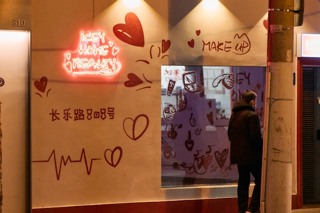 A storefront with a neon sign reading 'ICEY HOME BEAUTY' is illuminated. The wall features heart designs and various beauty-related doodles such as makeup items. A person stands outside the shop, leaning against a streetlight pole. The scene appears to be in an urban setting during the evening.