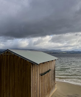 A wooden shack with a metal roof stands on a sandy shore next to a body of water. The sky above is filled with dark, heavy clouds, and in the distance, hilly terrain can be seen.