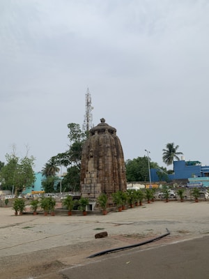 A small ancient stone temple stands prominently, surrounded by potted plants. The temple features intricate carvings on a cylindrical structure with a dome-shaped top. In the background, there are tall trees, a telecommunications tower, and modern buildings painted in blue. The area appears to be a mix of urban and historical elements.