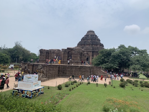 A large ancient stone temple with intricate carvings set amidst greenery. Numerous visitors are exploring the temple's exterior and walking around the gardened area. A signboard in the foreground promotes cultural heritage.