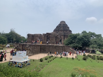 A large ancient stone temple with intricate carvings set amidst greenery. Numerous visitors are exploring the temple's exterior and walking around the gardened area. A signboard in the foreground promotes cultural heritage.