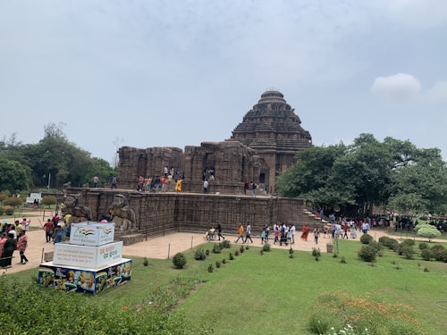 A large ancient stone temple with intricate carvings set amidst greenery. Numerous visitors are exploring the temple's exterior and walking around the gardened area. A signboard in the foreground promotes cultural heritage.