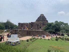 A large ancient stone temple with intricate carvings set amidst greenery. Numerous visitors are exploring the temple's exterior and walking around the gardened area. A signboard in the foreground promotes cultural heritage.