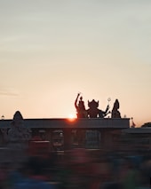 Silhouetted statues of Hindu deities are seen against the backdrop of a setting sun, creating a spiritual and serene atmosphere. The sky is clear with a gradient of colors from the setting sunlight.