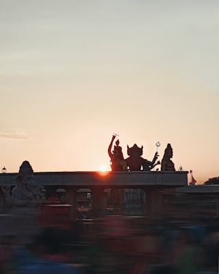 Silhouetted statues of Hindu deities are seen against the backdrop of a setting sun, creating a spiritual and serene atmosphere. The sky is clear with a gradient of colors from the setting sunlight.