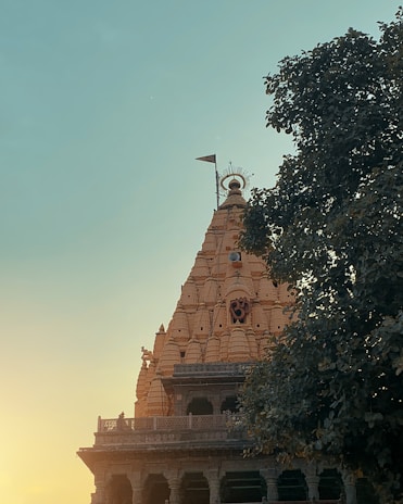 A majestic temple spire with intricate carvings rises into the clear sky, with a sacred symbol adorning its facade. A single flag atop the spire adds a sense of reverence. The warm sunlight bathes the temple in a gentle glow, contrasting with the deep green of the surrounding tree foliage.